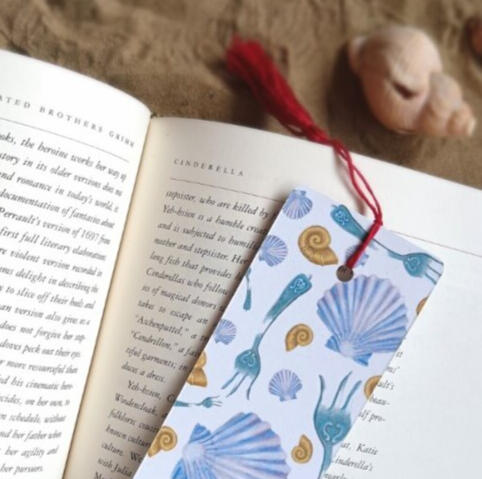 A beautiful bookmark covered in hand painted seashells and ornate forks lays on a book in the sand with a red handmade tassel.
